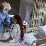 A female nursing assistant wearing full personal protective equipment stands at a patient’s bedside, adjusting an IV line. The patient is sitting up in her bed, wearing a hospital gown. Other hospital equipment is throughout the room.