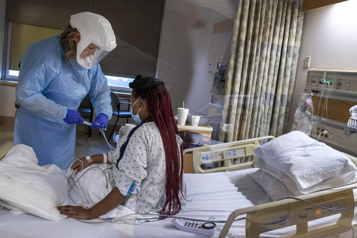 A female nursing assistant wearing full personal protective equipment stands at a patient’s bedside, adjusting an IV line. The patient is sitting up in her bed, wearing a hospital gown. Other hospital equipment is throughout the room.