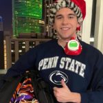 First-year College of Medicine medical student Marc Levine stands in front of a window at night wearing a Penn State T-shirt and Santa cap. He is giving the thumbs up and smiling. He has a face mask hanging from his neck and is holding a large bag with many face masks inside.