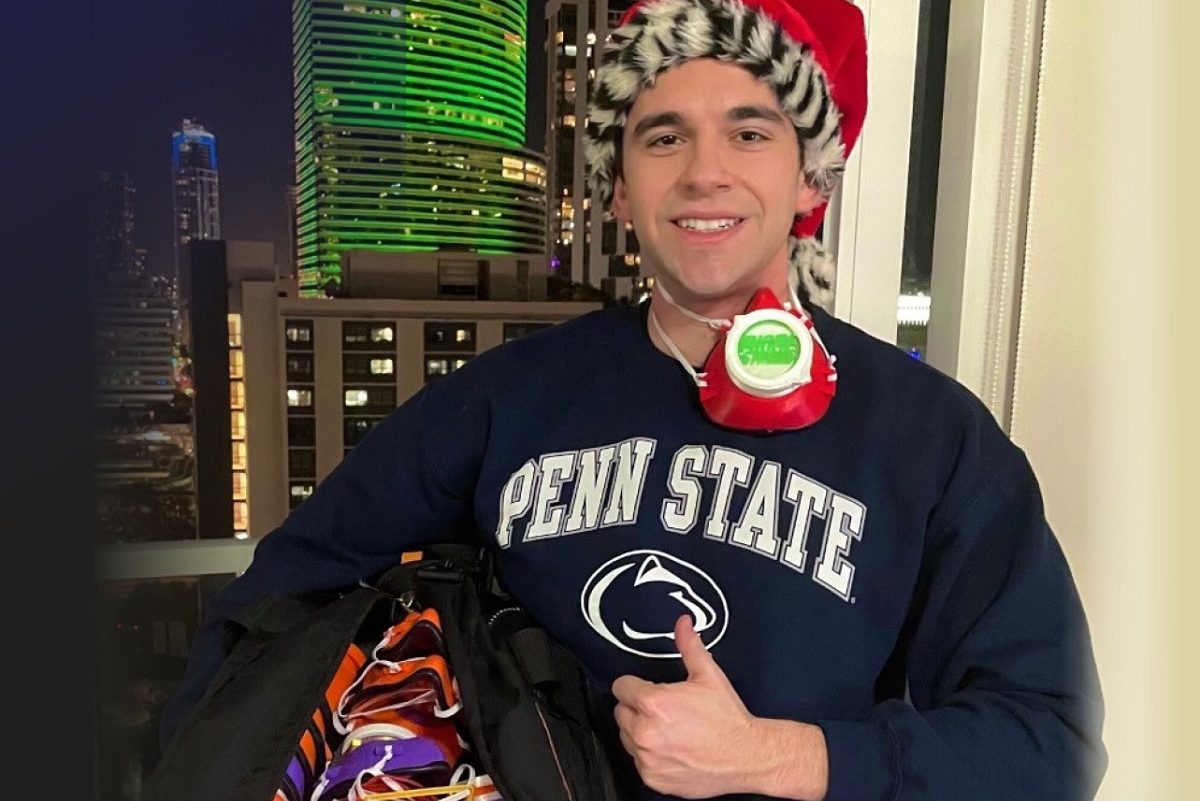 First-year College of Medicine medical student Marc Levine stands in front of a window at night wearing a Penn State T-shirt and Santa cap. He is giving the thumbs up and smiling. He has a face mask hanging from his neck and is holding a large bag with many face masks inside.