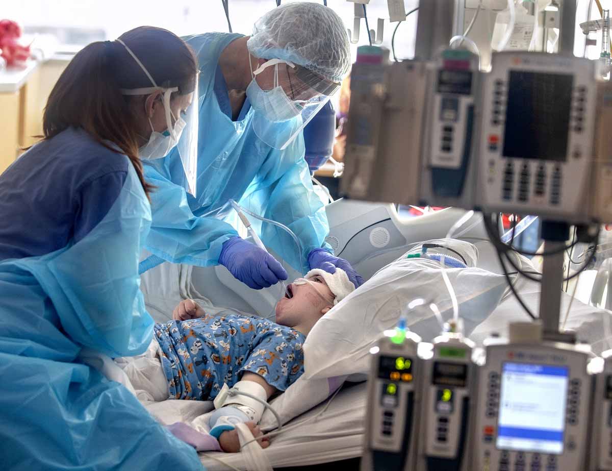 Dos trabajadores de la salud en equipo de protección personal deslizan un tubo por la garganta de un niño, mientras el niño descansa en una cama de hospital.