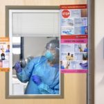 A medical professional in personal protective equipment gestures through the window in a door. Pasted to the door are posters for “contact precautions” and “airborne precautions.”