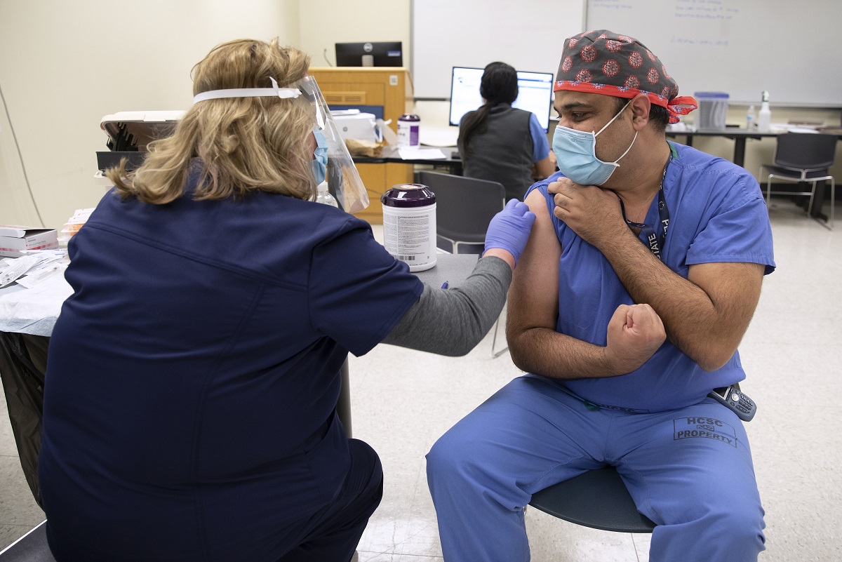 A man in scrubs, a head covering and a surgical masks, clenches his fist, while a woman in personal protective equipment prepares to give him a shot.