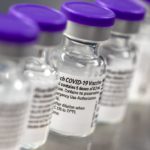 A row of vials show labels indicating they contain the COVID-19 vaccine.