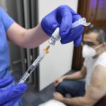 Hands in rubber gloves coming from a surgical down prepare a syringe of COVID-19 vaccine. In the background, Dr. Mohammad Ali sits in a short-sleeve shirt and a surgical mask.