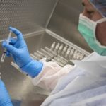 A woman in personal protective equipment inserts a syringe into a vial.