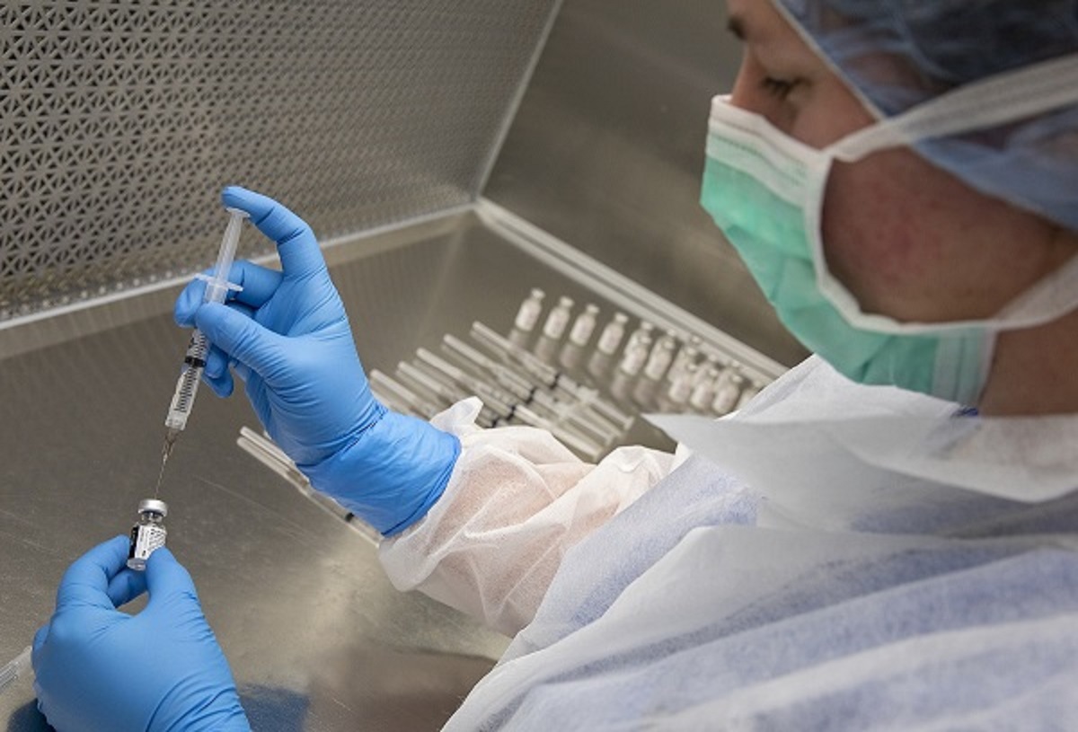 A woman in personal protective equipment inserts a syringe into a vial.