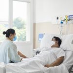 A woman in a mask sits at the end of a hospital bed, in which lies a man a man in a mask.