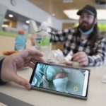 A hand holds a smart phone upon which is the image of an infant in a bed. Across a lunch table littered with bottles and food, a man with a beard and a hat looks on out of focus.