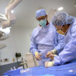 Two medical professionals wearing blue scrubs, facemasks and gloves perform a biopsy on a heart transplant patient at Penn State Health Milton S. Hershey Medical Center.