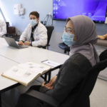 In the foreground, a female medical student wearing a surgical mask and head covering is seated at a conference table with an open textbook in front of her. In the background, two male students, also wearing surgical masks, are seated at adjoining conference tables with open laptops in front of them.
