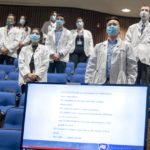 Ten students wearing white physician coats and masks stand in front of blue seats in Junker Auditorium. A large computer screen with details of the Student Clinician Ceremonies is set in front of the student seating.