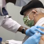 A man in a hat and a surgical mask receives an injection from a health care worker.