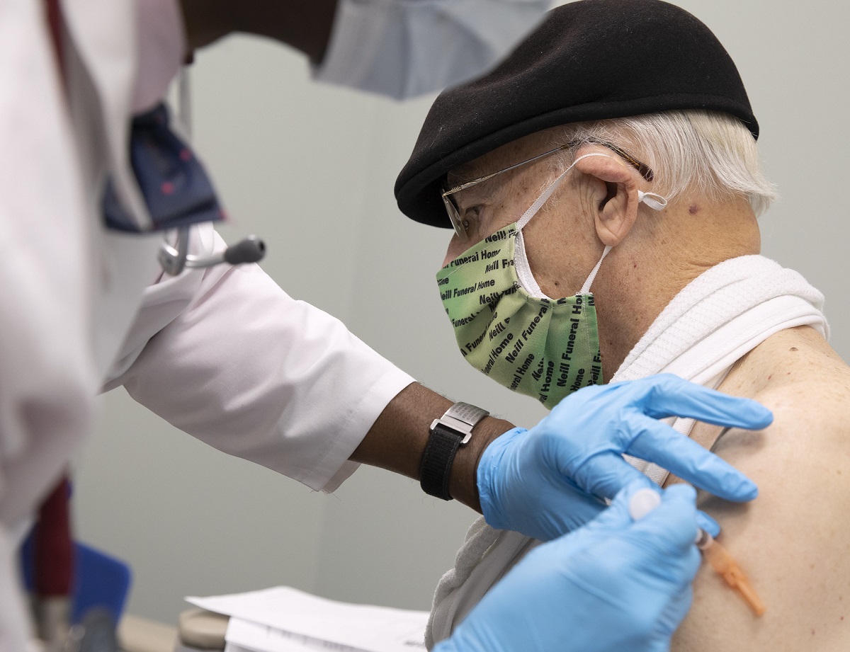 A man in a hat and a surgical mask receives an injection from a health care worker.