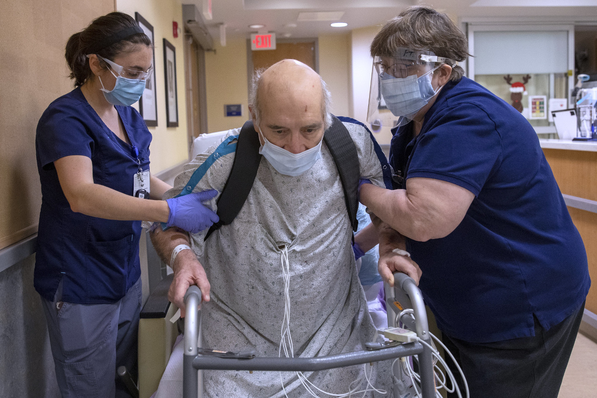 A man in a hospital gown rises out of a wheelchair to a standing position, leaning on a walker. Several wires emerge from his gown. Two staff members, one on each side, are bracing the man’s upper arms.