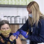 An older woman, wearing a face mask and with her eye glasses propped up on her head, is seated, with her short sleeve pulled up to her shoulder. A health care worker, wearing blue surgical gloves and a mask, stands beside her to administer a shot in her upper arm.