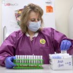 Angie Spohn stands in front of two large refrigerators in the lab and transfers test tubes from a rack into box. She wears a mask, lab coat and a pin that says ‘I got my COVID-19 vaccine!’