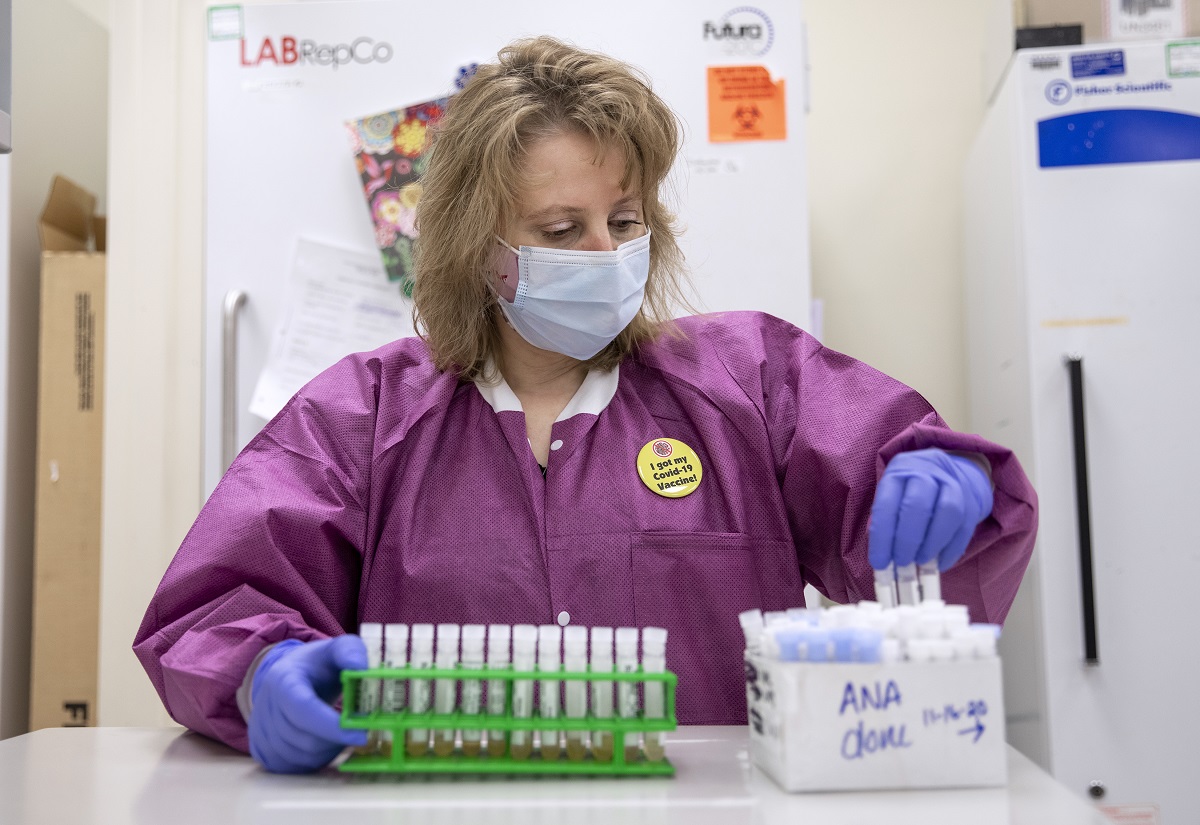 Angie Spohn stands in front of two large refrigerators in the lab and transfers test tubes from a rack into box. She wears a mask, lab coat and a pin that says ‘I got my COVID-19 vaccine!’