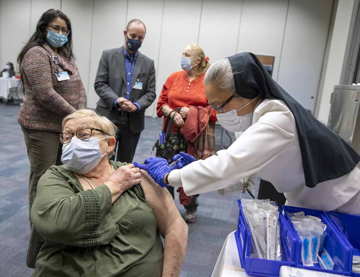 Una mujer con una máscara mira hacia otro lado mientras otra mujer con ropa del hospital, guantes de goma y el hábito de una monja le da una inyección en el hombro.