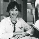 Mary Simmonds sits at a desk wearing a lab coat and stethoscope