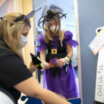 A young girl in a Halloween dress and spider hat stands in the doorway of a hospital room while a woman kneels down near her, holding up a doll-like toy.