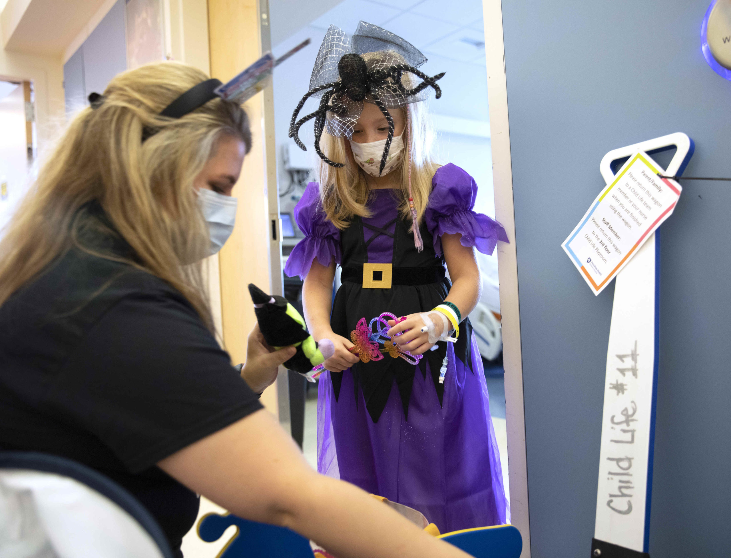 A young girl in a Halloween dress and spider hat stands in the doorway of a hospital room while a woman kneels down near her, holding up a doll-like toy.