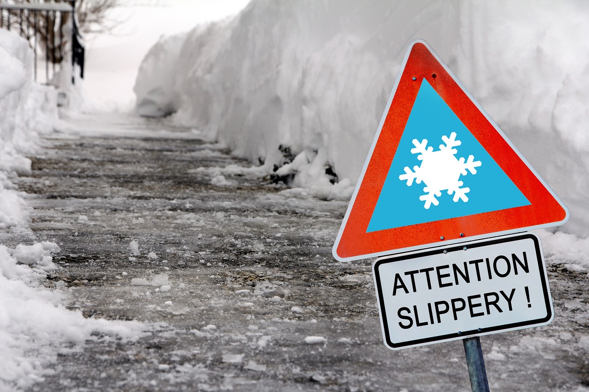 An icy sidewalk in winter with a sign in the foreground reading ‘Attention slippery!’