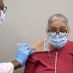 A male health care provider, left side of photo, wearing glasses, a face mask and blue medical gloves, prepares to inject the COVID-19 vaccine into the upper arm of an elderly woman who is wearing a face mask.
