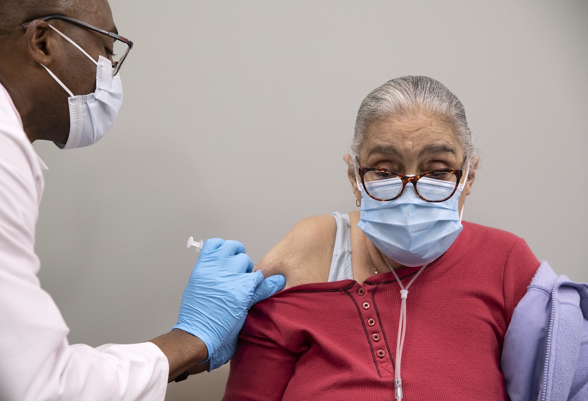 A male health care provider, left side of photo, wearing glasses, a face mask and blue medical gloves, prepares to inject the COVID-19 vaccine into the upper arm of an elderly woman who is wearing a face mask.