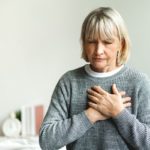 A woman sits on the edge of a bed with chest pain, experiencing a heart attack.