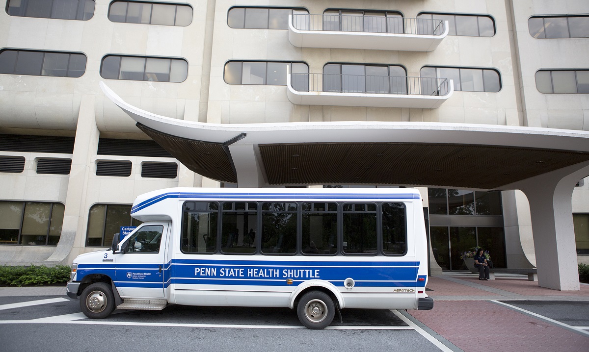 Bus with the words Penn State Health Shuttle printed on the side parked outside a hospital entrance.