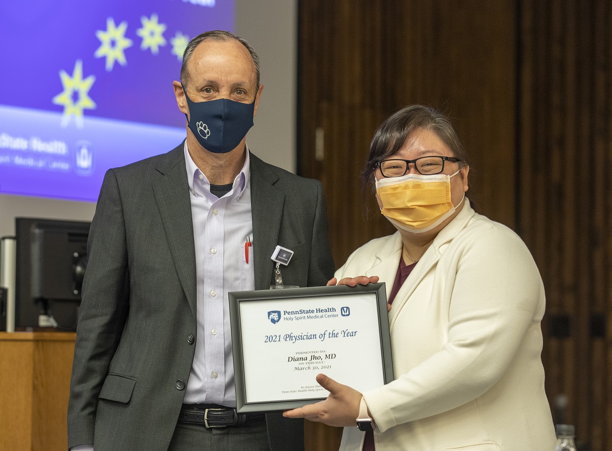 A man dressed in a gray suit and white shirt and wearing a navy blue face mask stands with a woman dressed in a while coat and dark blouse, and wearing glasses and an orange face mask, while holding a framed award.