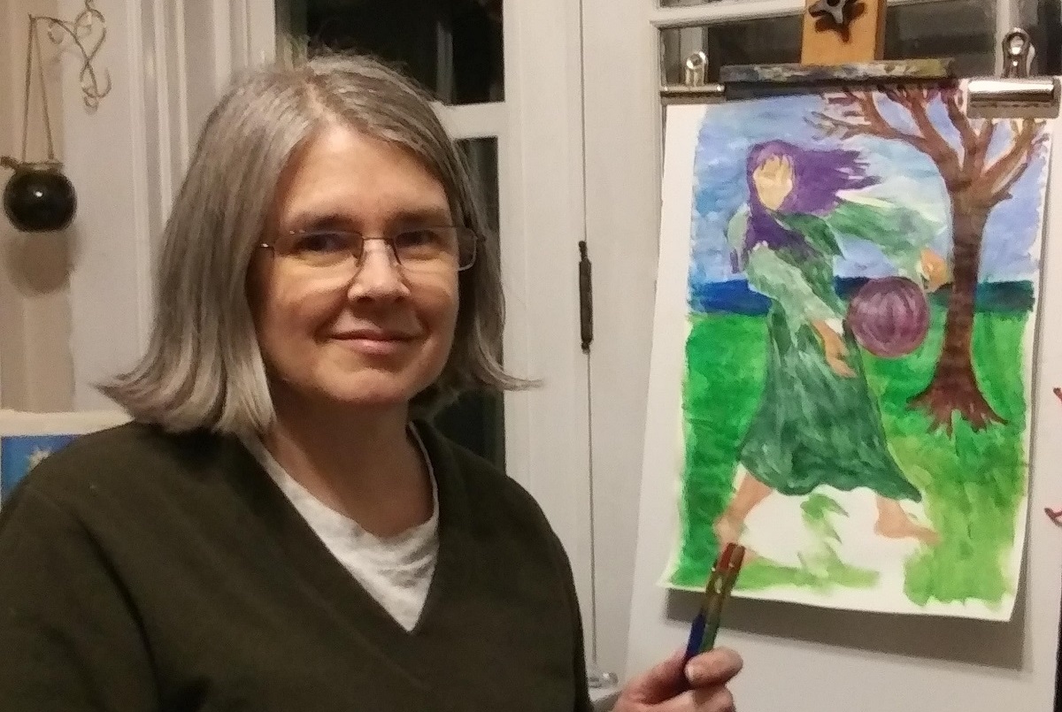 Jennifer Rudolph, who wears glasses and a V-neck sweater, sits at an easel and holds a brush in her hand. A painting on the easel shows a girl standing by a tree with her hair blowing in the wind.