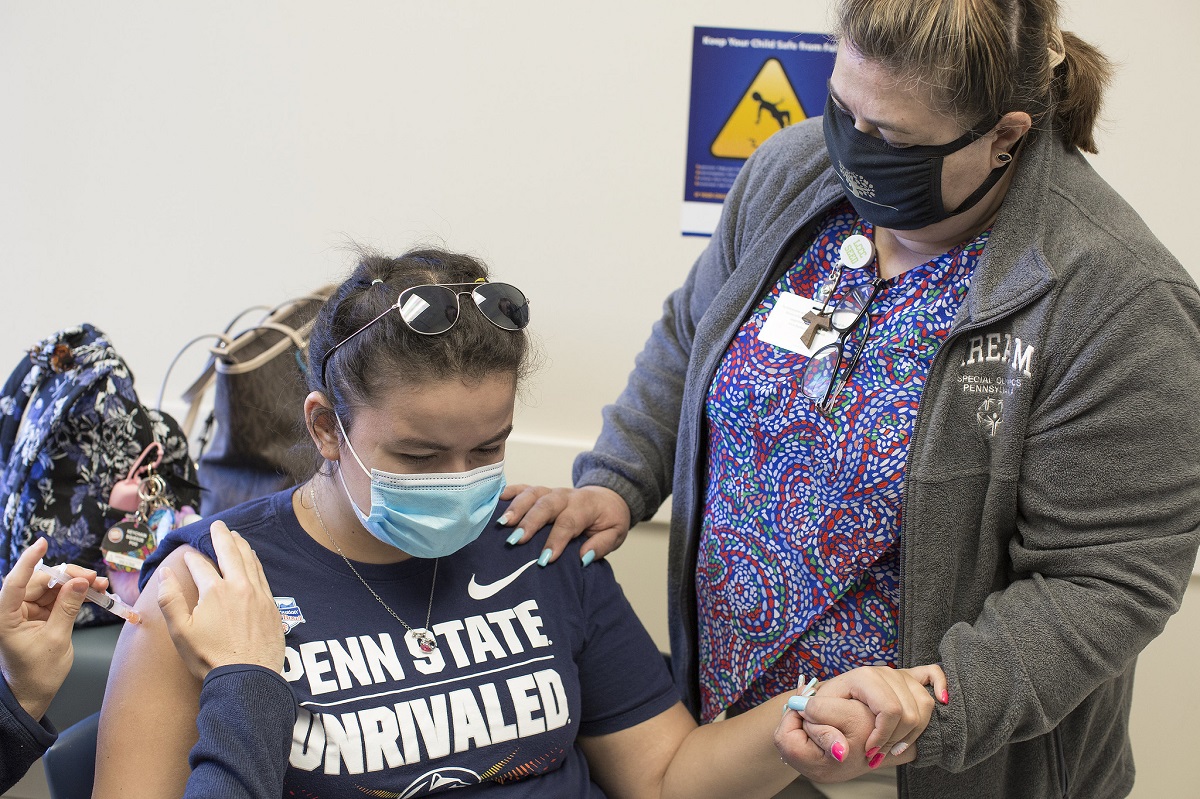 Rachel Frometa, who is seated and wears a mask and sunglasses on the top of her head, holds the hand of her mother, Betty Frometa, who stands beside her, as a vaccine is pressed into her arm by the hands of a health care worker.