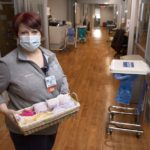 A woman in a mask stands in a clinic hallway with stained-wood floors holding a basket of feminine hygiene products.