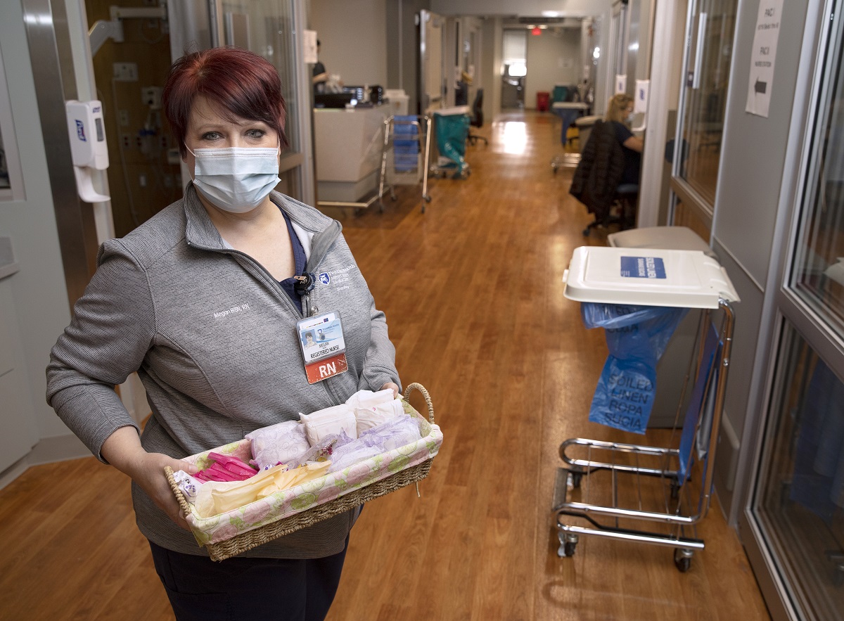 A woman in a mask stands in a clinic hallway with stained-wood floors holding a basket of feminine hygiene products.