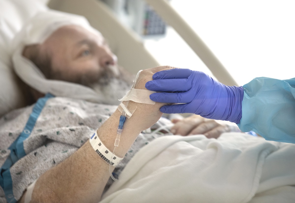 A male patient wearing a white and blue hospital gown reclines in a hospital bed. A nurse’s hand, covered in a blue glove, grasps his hand, helping him to move.