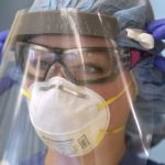 A close-up of a nurse putting on a face shield over an N-95 mask and goggles. She is also wearing a hair covering and surgical gloves.
