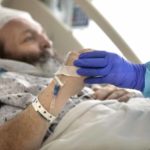 Un paciente masculino con una bata de hospital blanca y azul se reclina en una cama de hospital. La mano de una enfermera, cubierta con un guante azul, agarra su mano, ayudándole a moverse.