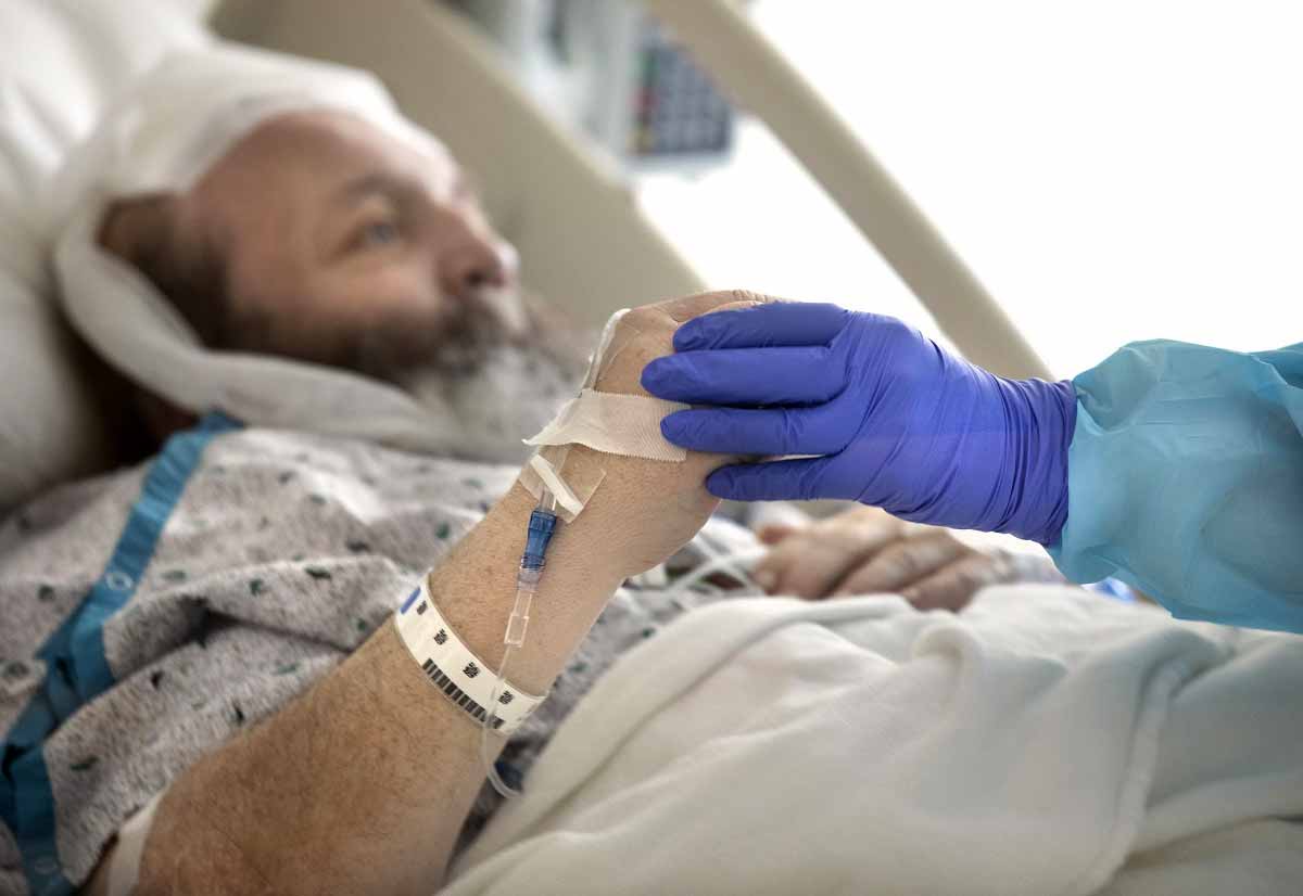 Un paciente masculino con una bata de hospital blanca y azul se reclina en una cama de hospital. La mano de una enfermera, cubierta con un guante azul, agarra su mano, ayudándole a moverse.