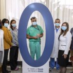 A group of six people stand on either side of a large cardboard cutout of the numeral zero at the 2019/2020 Patient Safety & Reliability awards event in October 2020. A man dressed in scrubs stands in the middle of the zero.