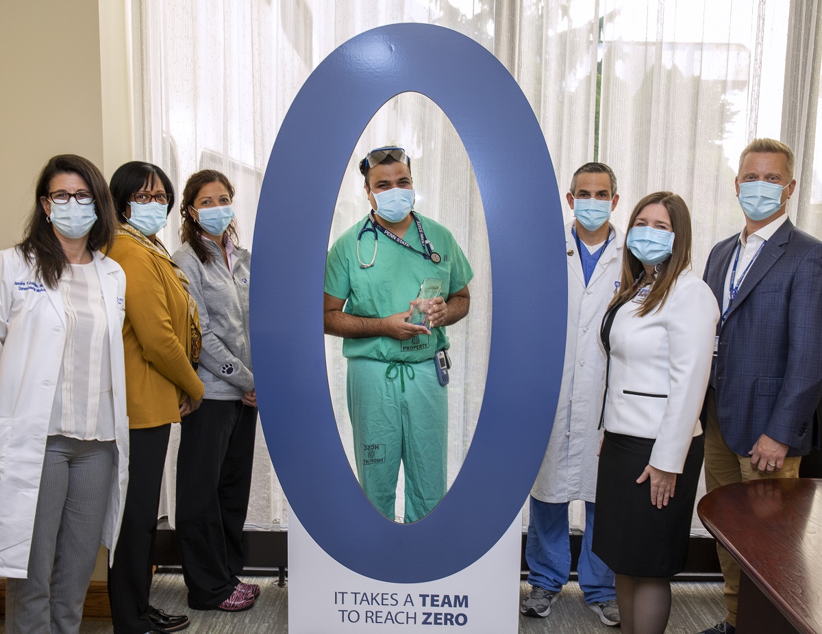 A group of six people stand on either side of a large cardboard cutout of the numeral zero at the 2019/2020 Patient Safety & Reliability awards event in October 2020. A man dressed in scrubs stands in the middle of the zero.