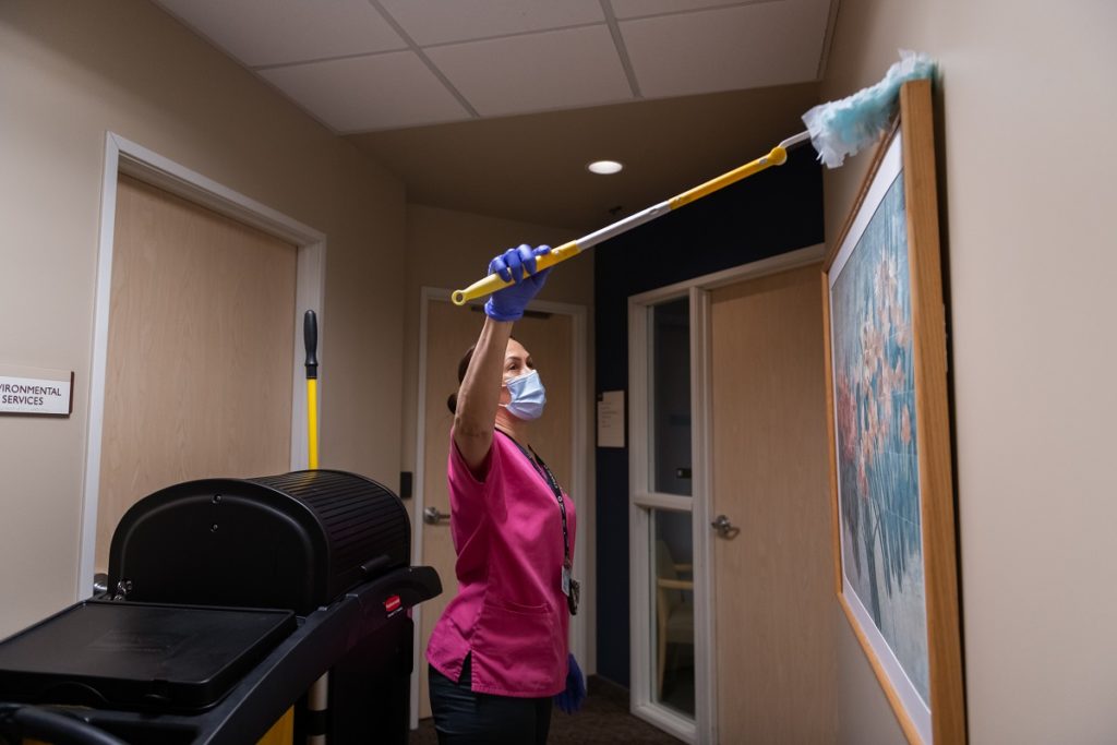Marisol Rosa, an environmental services associate with St. Joseph Medical Center, holds a long-handled duster and dusts a painting. She is wearing a smock, a lanyard with a nametag and a facemask. Behind her is a cleaning supplies cart.