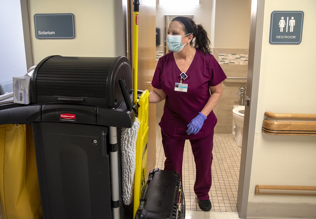 Dianne Finucane, an EHS associate with Holy Spirit Medical Center, smiles as she walks out of a bathroom. She is wearing a smock, pants, a face mask and has a nametag clipped to her blouse.