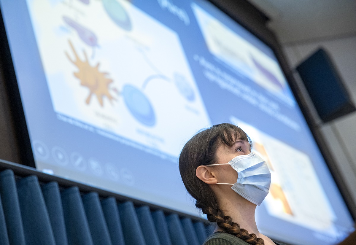 Elizabeth Lesko is seen in profile in front of a large screen with a PowerPoint slide on it. She is wearing a face mask and has her hair in a braid.