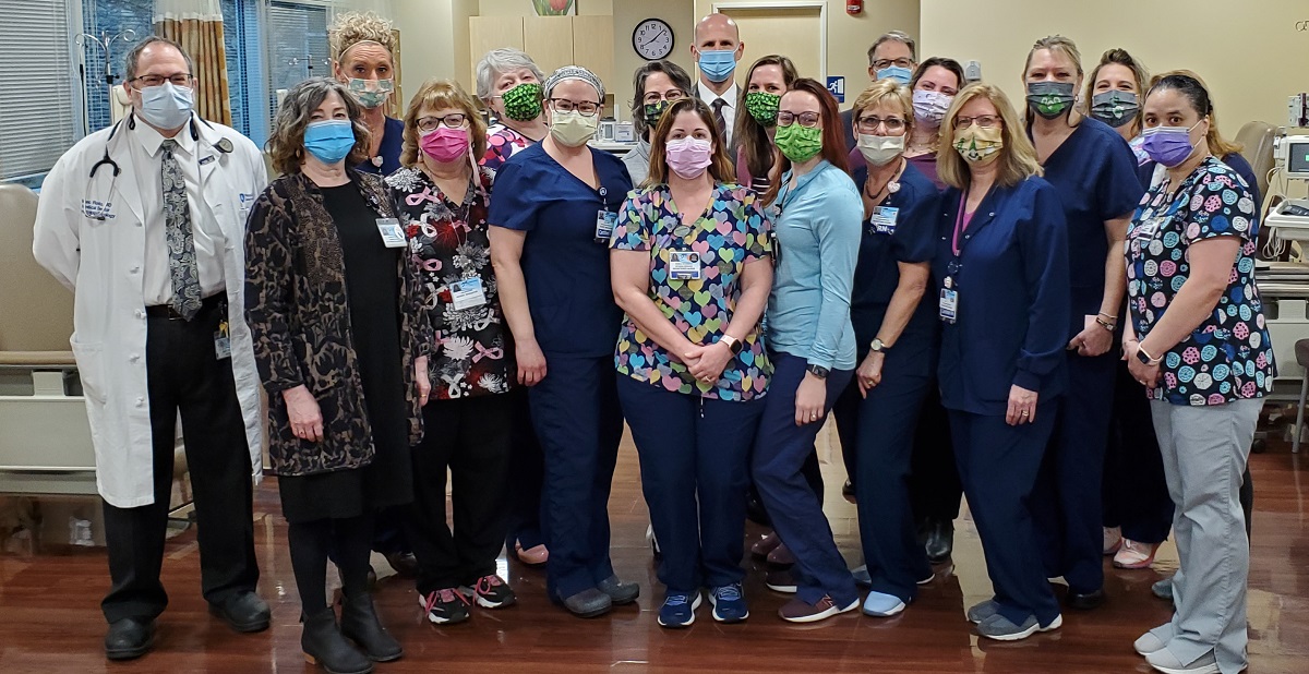 A group of health care workers in scrubs stand in a hospital room and pose for a photo.