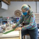 Linda Billet perches on a stool in her workshop and draws with a marker on a strip of glass. She wears glasses and a face mask and has her hair in a ponytail streaked with colors. Art supplies surround her on the counter where she works.