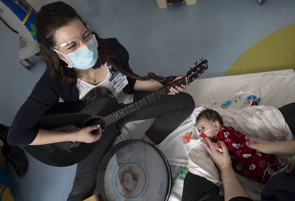 Tina Myers, who wears a mask and goggles, sits on the edge of a mattress on the floor and plays her guitar for a baby who is lying on the mattress. The baby, who sucks on a pacifier, is wearing a sleeper with snowmen on it and is held by a pair of hands.