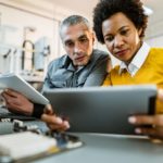 A man and a woman look at a computer tablet. The man is holding an open book and has his hands resting on a keyboard.