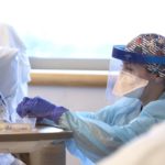 An elderly woman with glasses is sitting up in her hospital bed, food tray in front of her. A female nurse wearing a face mask, a face shield, blue surgical gloves and blue protective gown, crouches by the bed to place medicine in a cup.